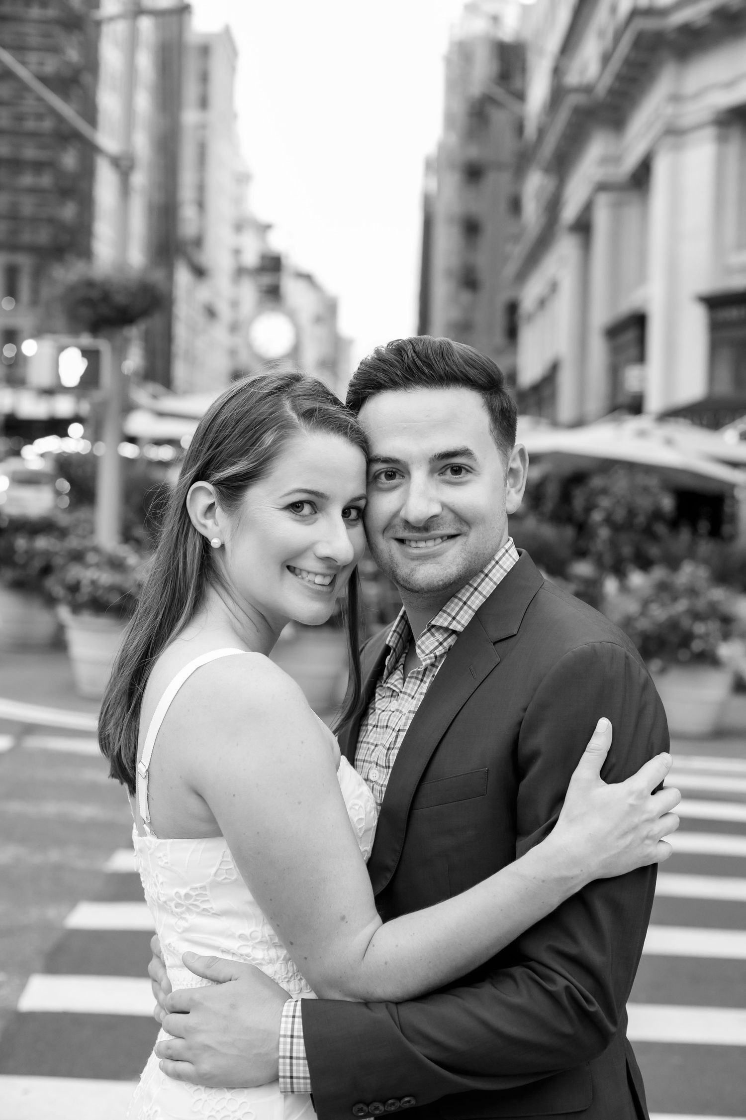 A romantic black and white portrait of a couple embracing on a city street with urban buildings in the background.