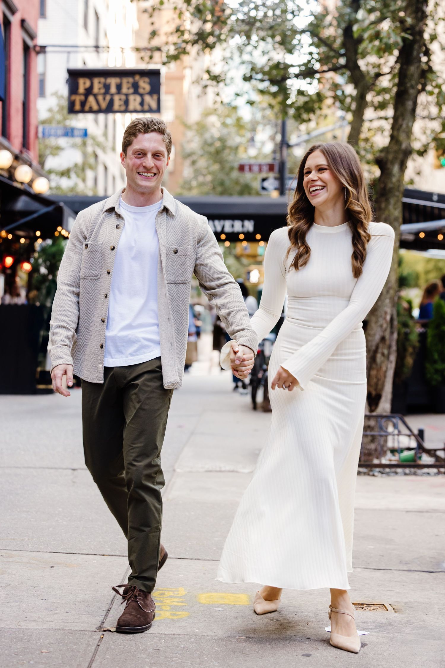 A couple strolls together on a city sidewalk, wearing coordinated white and beige outfits on a sunny day.