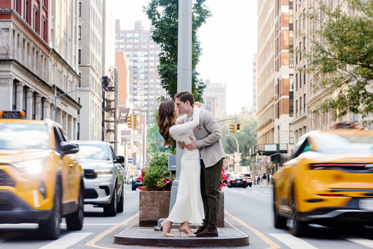 A couple embraces on a city sidewalk with yellow taxis and tall buildings visible in the urban background.