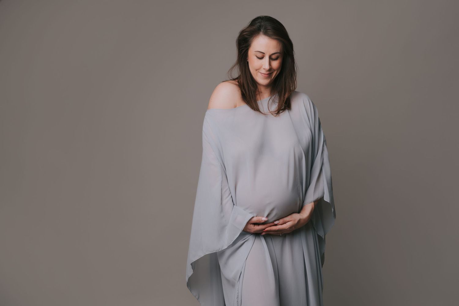 Woman in flowing gray dress gently cradles her pregnant belly against neutral backdrop during maternity photoshoot.