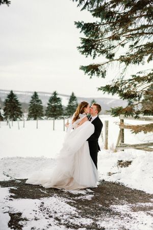 A romantic winter wedding scene with a couple embracing on a snowy path surrounded by evergreen trees in the mountains.