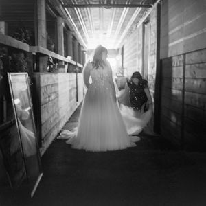 A silhouetted figure in a flowing wedding dress stands in a sunlit barn corridor with dramatic backlighting.