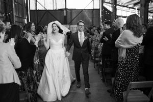 A monochrome wedding photo showing a couple walking down an aisle surrounded by guests at an indoor venue.