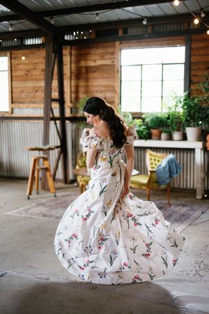 A woman in a flowing floral dress twirls gracefully in a rustic indoor setting with wooden walls and potted plants.