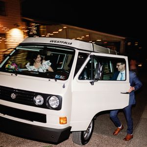 A white Volkswagen Westfalia van parked at night with doors open and warm lighting in the background.
