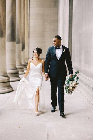 A bride in a white satin gown and groom in classic tuxedo walk together holding hands along a columned hallway.
