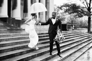 A couple in wedding attire shares a joyful moment under a white umbrella on rainy church steps in black and white.