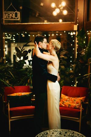 A romantic wedding couple shares an intimate kiss in a dimly lit venue with red velvet seating and twinkling lights.