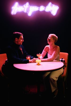 Two people sit across from each other at a round table in a dimly lit room with pink neon lighting above.