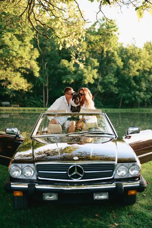 Couple with dog in vintage black Mercedes-Benz convertible surrounded by lush green trees near a pond.