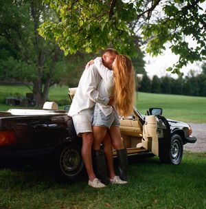 Couple kissing and embracing beside a vintage convertible car under a green tree on a sunny day.