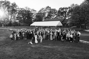 Large group gathered outdoors for wedding celebration in front of rustic barn building in black and white photograph.