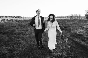 A happy newlywed couple walks together across a grassy hillside, captured in black and white photography.