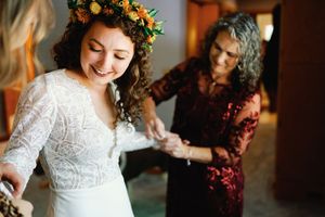 A joyful bride in a lace wedding dress and floral crown gets help preparing for her wedding ceremony.