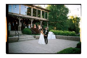 Bride and groom share a romantic outdoor dance at a rustic wedding venue with string lights and lush greenery.
