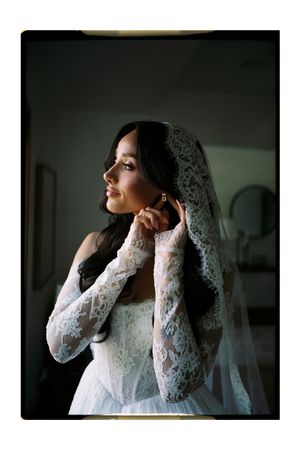 Bride in lace wedding dress and veil adjusts earring by window light.
