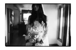 Black and white photo of a bride in a lace gown holding a floral bouquet in a softly lit hallway.