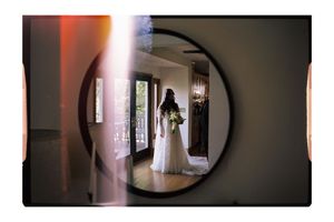Bride in white gown holding bouquet reflected in circular mirror during wedding preparation.
