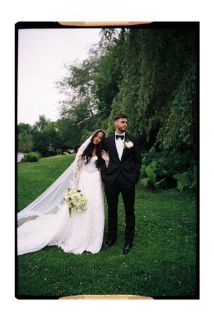 A bride in a white gown with flowing veil holds bouquet beside a groom in black tuxedo on lush green lawn.