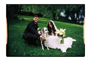 Bride and groom sitting on green grass with a Dalmatian dog, bride holding white bouquet, outdoor wedding photo.