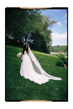 Bride in white lace gown with long veil holds bouquet on green lawn with trees and blue sky.