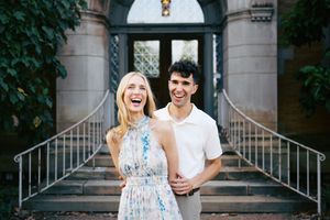 Happy couple laughing together in front of elegant stone building with iron railings on a sunny day.