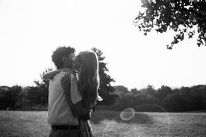 Black and white photo of a couple embracing and kissing in a park with trees in the background.