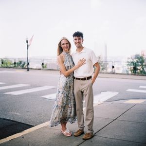 A smiling couple poses on a sunny sidewalk near a crosswalk, woman in floral dress, man in white shirt and khakis.