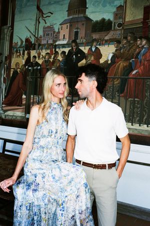 Elegant couple posing before a Renaissance mural painting, woman in floral maxi dress, man in white polo shirt.