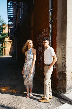 A woman in a floral midi dress and a man in white shirt and khaki pants pose in a urban alley with brick walls.