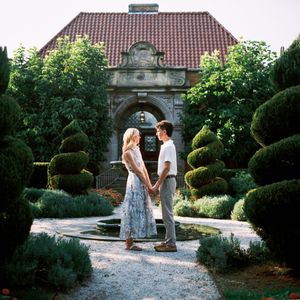 Couple holding hands in a formal garden with sculpted topiaries and a stone archway building backdrop.