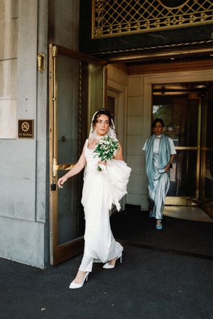 A bride in a flowing white gown and veil exits an ornate building entrance while holding a bouquet.