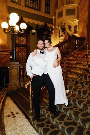 A romantic couple in formal attire poses on an elegant vintage staircase with ornate railings and classic lighting.