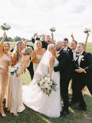 Wedding party in white dresses and black suits poses joyfully together outdoors on a sunny day.