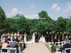 Outdoor wedding ceremony setup with white chairs and floral arch surrounded by lush green trees on a bright sunny day.
