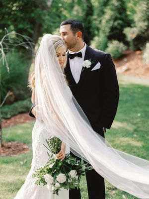 Classic wedding couple in black and white attire poses outdoors with a flowing cathedral veil in multiple romantic shots.