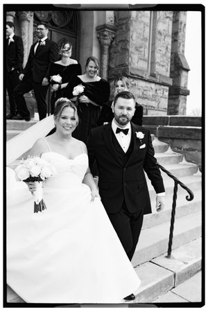 A joyful couple in formal wedding attire share a laughing moment together on stone steps outside a church.