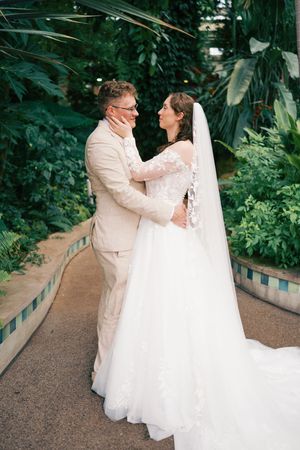 A bride and groom embrace in a lush botanical garden, she wearing a white lace gown with veil, he in a beige suit.