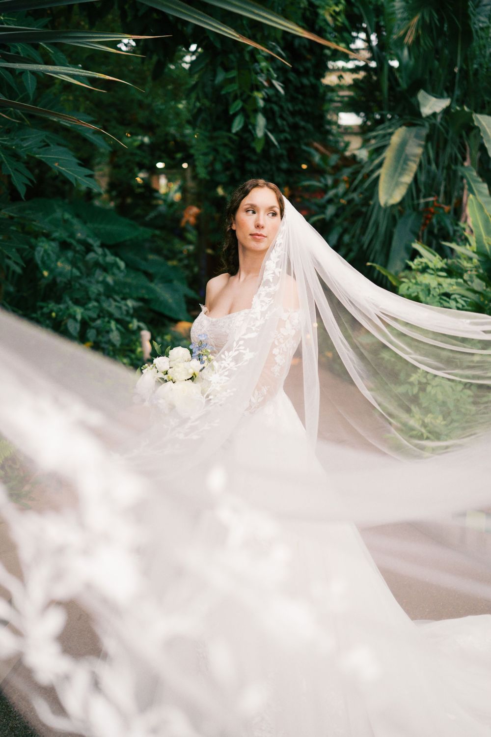 Bride in lace wedding gown and flowing veil holds white bouquet surrounded by lush tropical greenery.