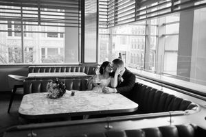 A couple shares an intimate moment at a diner booth next to large windows in black and white photography.