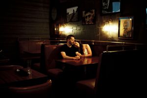 A man sits alone in a dimly lit bar booth, lost in thought, with warm lamp light and artwork on dark walls.