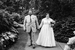 Black and white photo of a bride and groom holding hands, walking along a garden path lined with lush greenery.