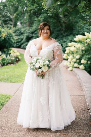 Plus-size bride in lace off-shoulder gown holds white bouquet on garden path surrounded by lush greenery.