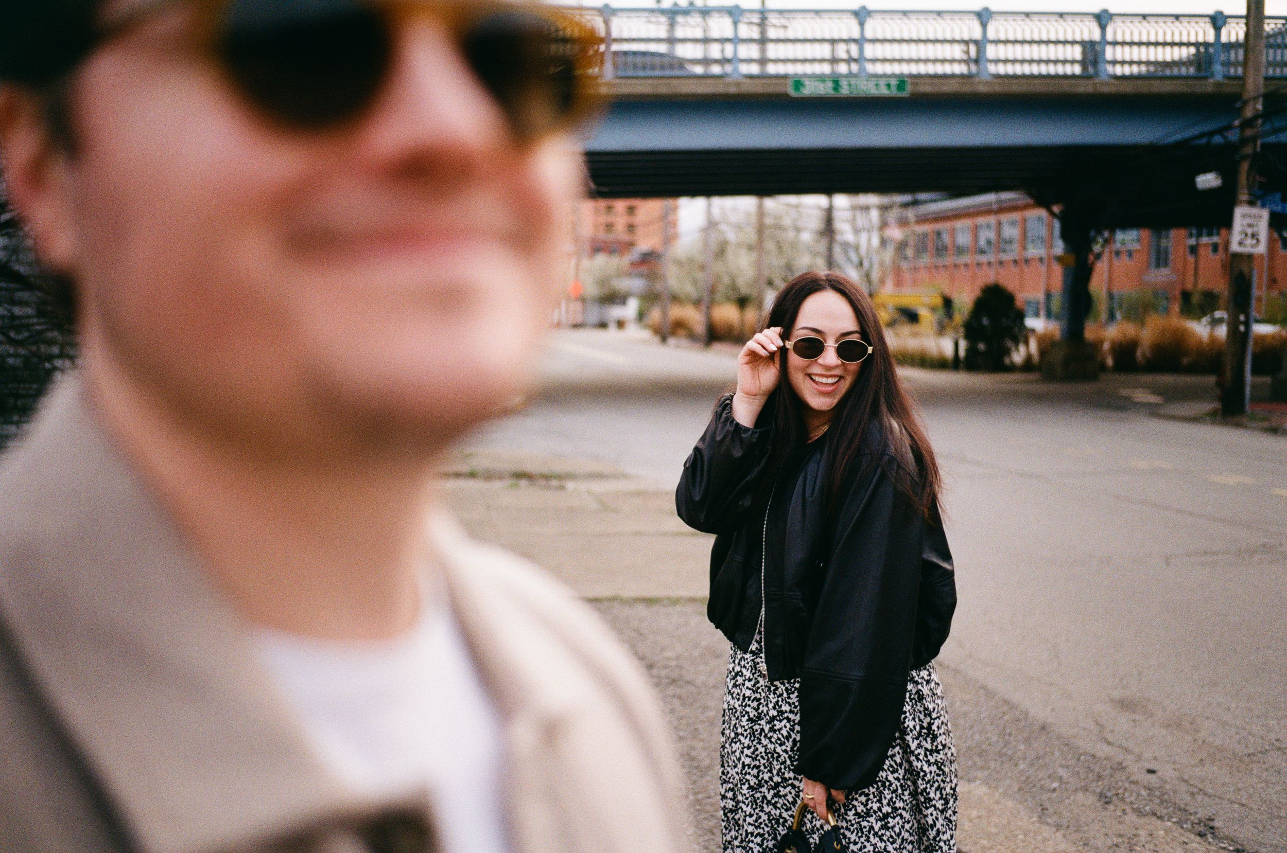 Two people wearing sunglasses pose casually on an urban street with a bridge overpass visible in the background.