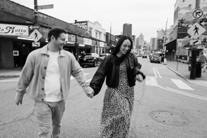 Black and white photo showing a couple holding hands and walking down a city street with storefronts and traffic signs visible.