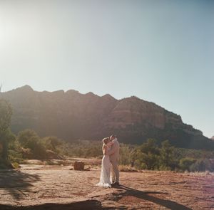 A couple stands on red desert rocks with towering mountain cliffs in the background during golden hour sunset.