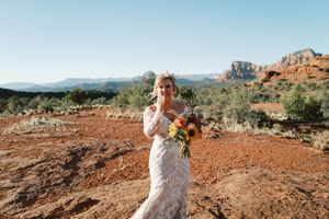 A person in a white lace wedding dress holds a sunflower bouquet on red desert soil with mountains in the background.