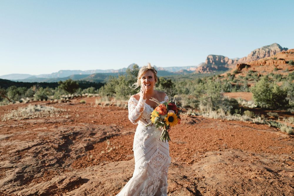 A person in a white lace wedding dress holds a sunflower bouquet on red desert soil with mountains in the background.