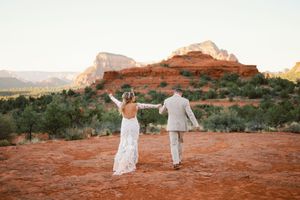 A couple in light colored wedding attire holds hands while walking on red rocks in Sedona, Arizona.