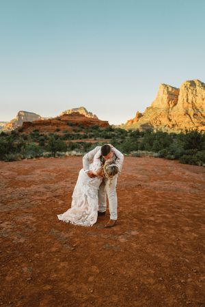 A romantic couple in wedding attire embrace against the stunning red rock formations of Sedona at sunset.